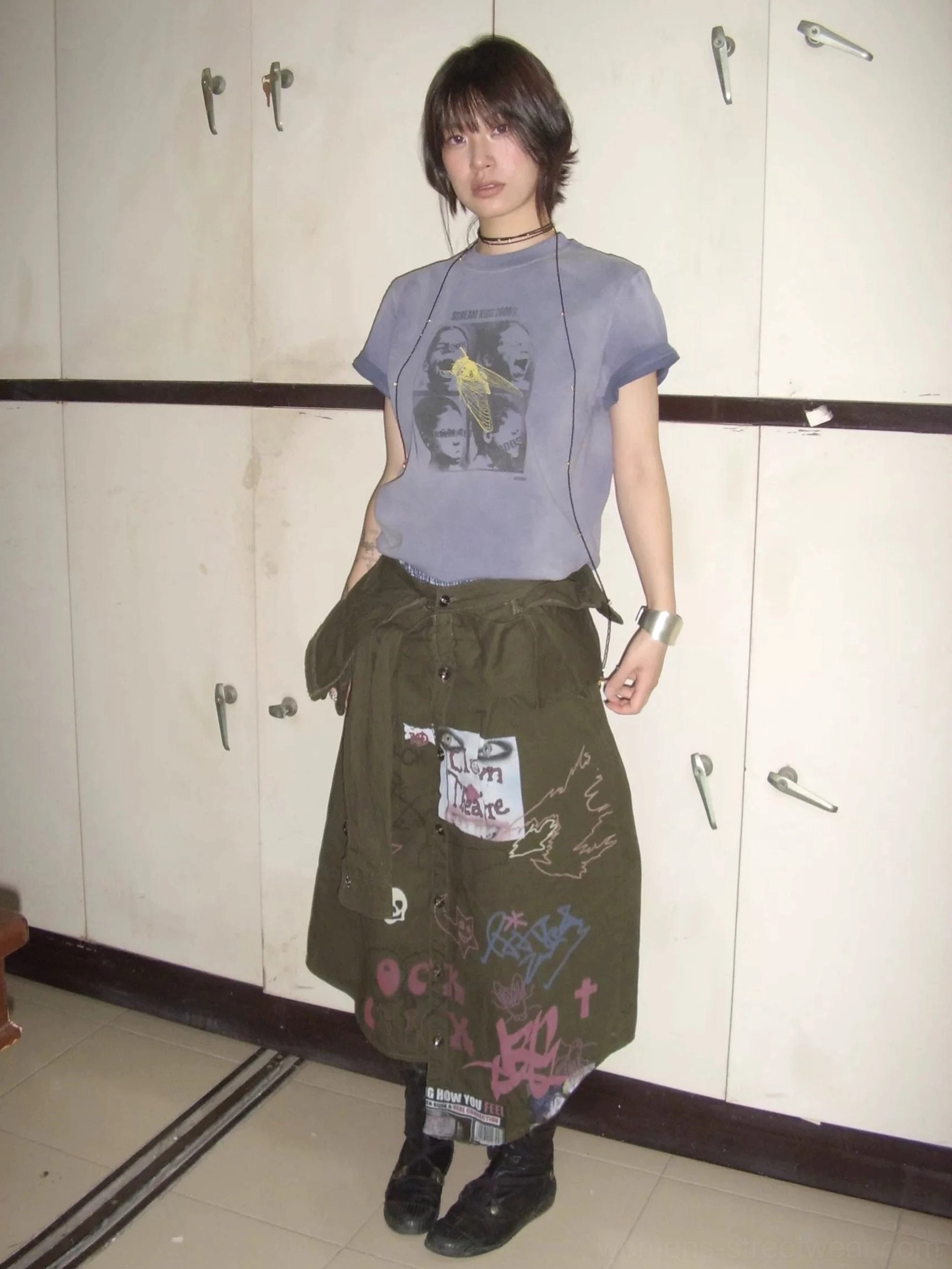 Full body female model wearing a washed purple blue Scream Kids graphic T-shirt styled with a printed cargo skirt in a locker room