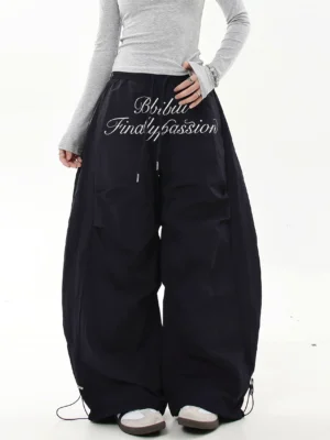 Black oversized baggy parachute cargo pants with white cursive script embroidery, adjustable waist and bungee ankle cuffs in a modern streetwear outfit