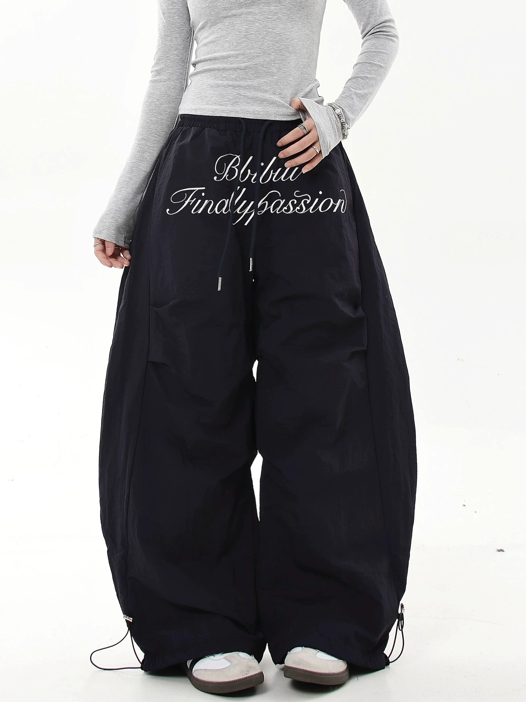 Black oversized baggy parachute cargo pants with white cursive script embroidery, adjustable waist and bungee ankle cuffs in a modern streetwear outfit