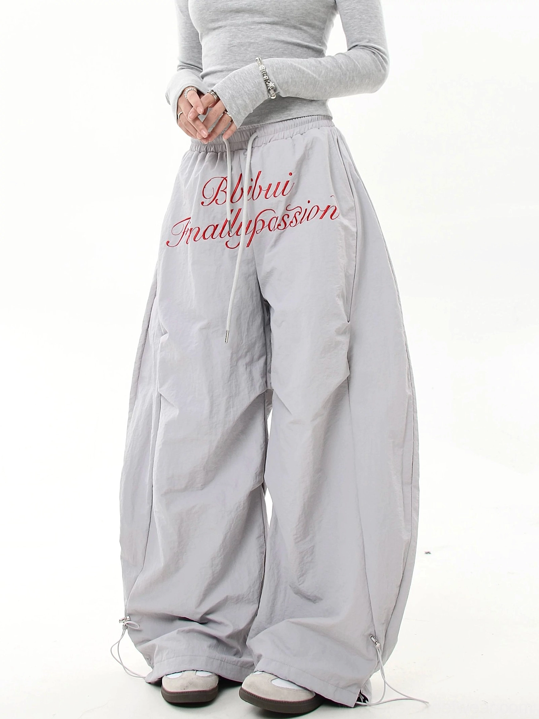 Light gray oversized parachute streetwear cargo pants with red script embroidery, adjustable drawstring waist and wide ankle cuffs styled with a fitted crop top