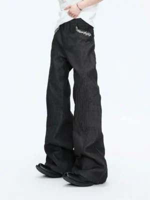Low angle front view of sculpted seam wide-leg black jeans with silver embroidered front pockets, dimensional leg pleats, vintage washed denim, white top, and black leather shoes