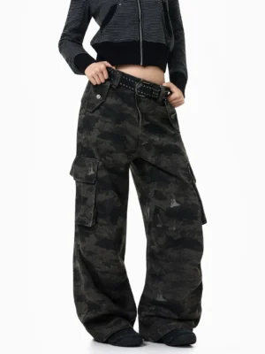 Front three-quarter view of dark camouflage baggy streetwear cargo pants styled with a striped cropped zip top, studded belt, and black sneakers