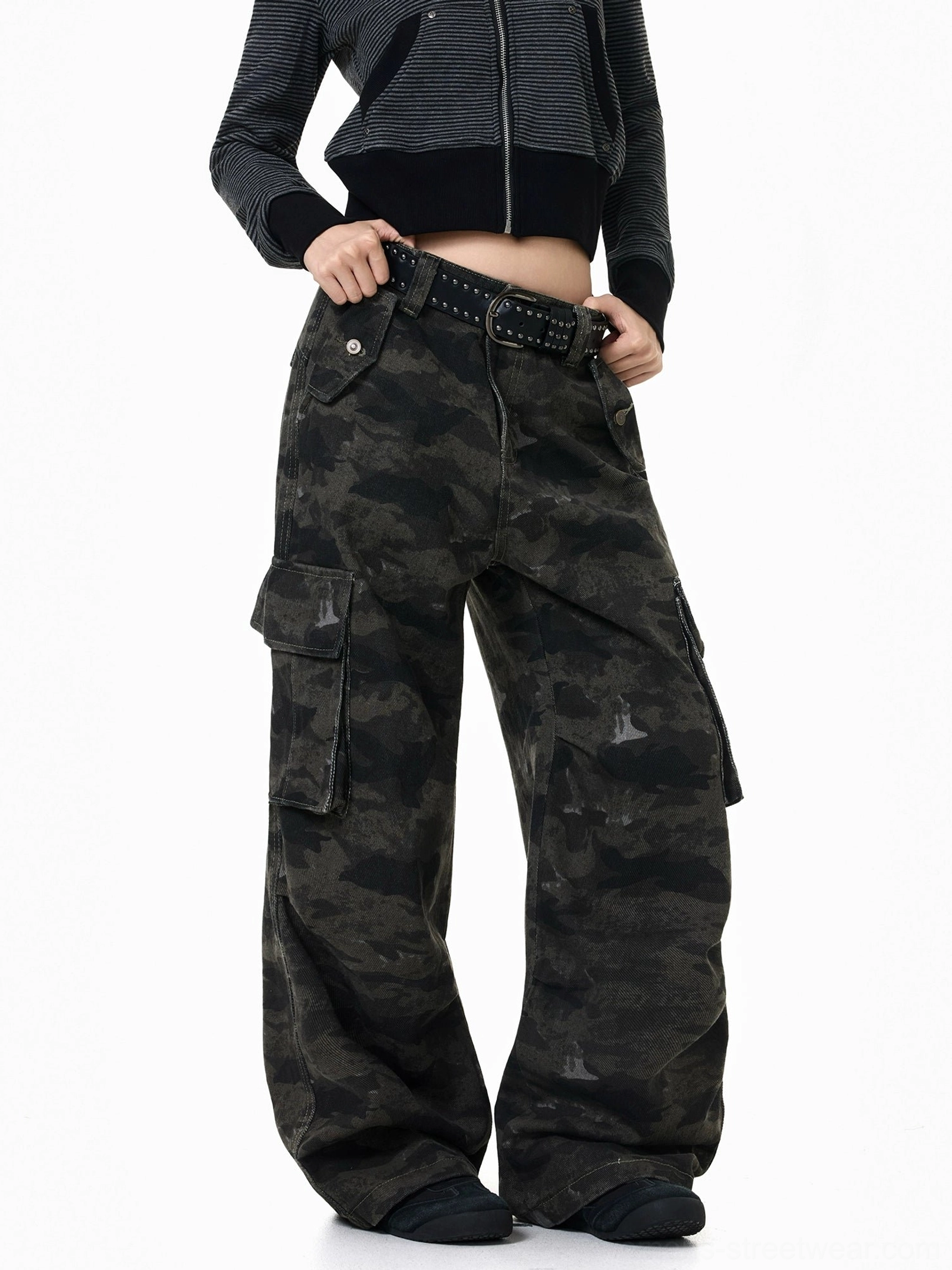 Front three-quarter view of dark camouflage baggy streetwear cargo pants styled with a striped cropped zip top, studded belt, and black sneakers