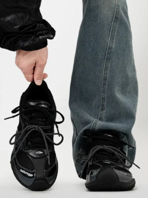 All-black Shadow Stream retro mesh running sneaker shown from a low front angle with a model wearing the shoe with baggy blue jeans, featuring layered mesh panels, synthetic overlays, flat laces, and subtle toe detailing