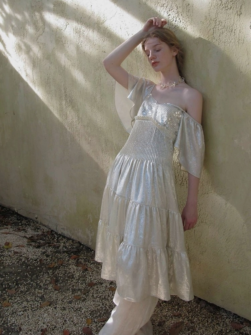 Rear three quarter view of champagne glossy off shoulder tiered dress with flared ruffle sleeves and layered skirt