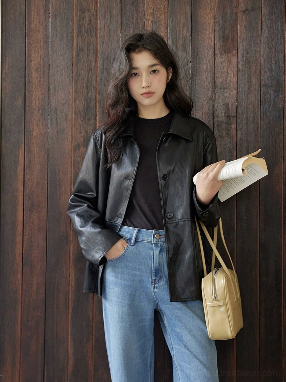 Full-body view of a woman wearing a short black leather jacket with blue jeans and a beige handbag