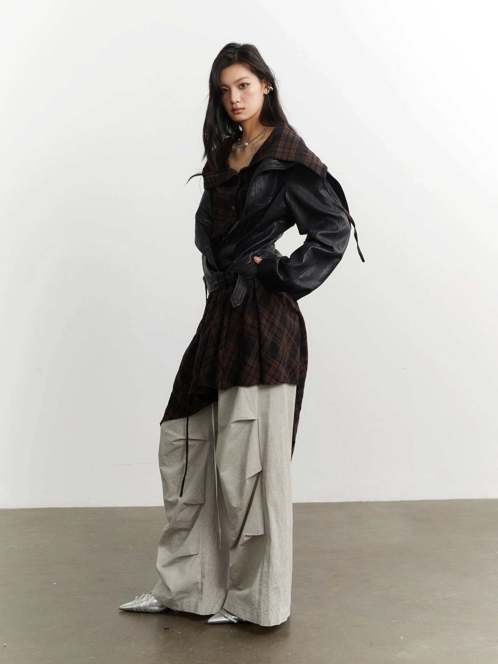 Front three-quarter view of a short-hem black faux leather jacket with asymmetric tartan plaid panel and contemporary streetwear cargo styling