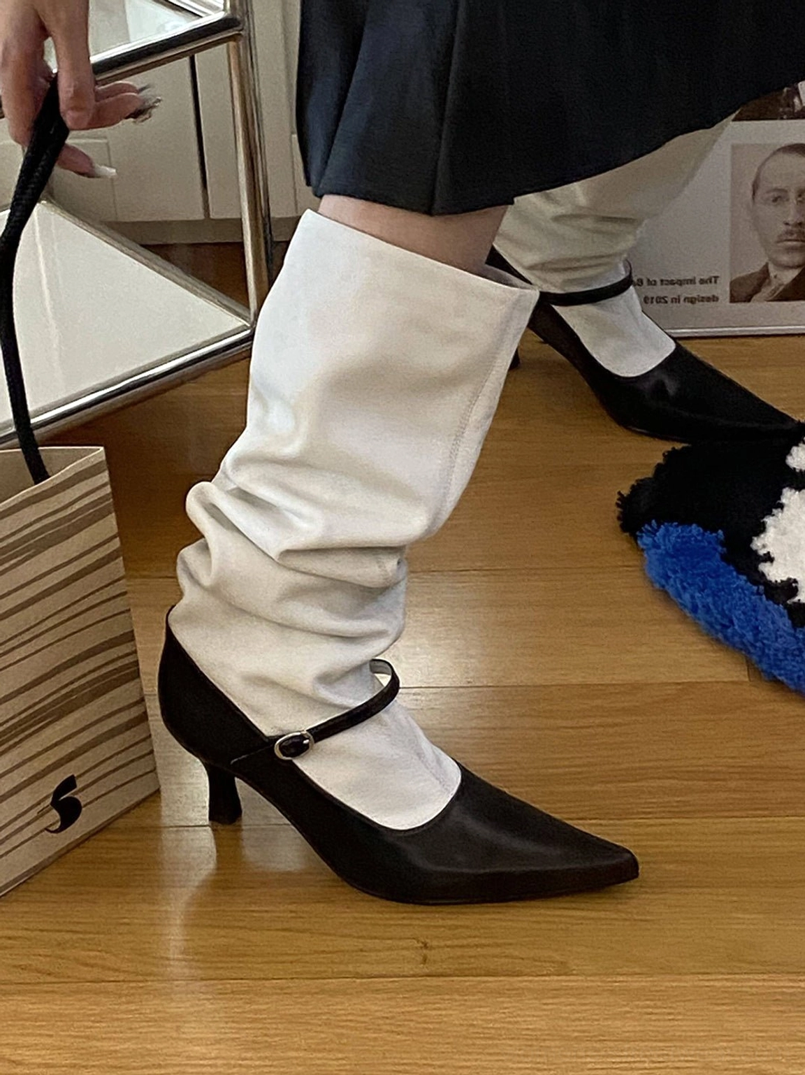 Side view of a model wearing black and white pointed-toe knee-high boots with gathered white shaft, black low stiletto heel, and adjustable ankle buckle