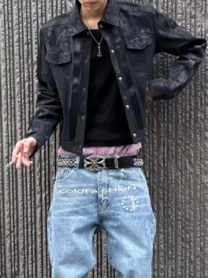 Male model wearing a silver-black faux leather trucker jacket with snap buttons, chest pockets, printed denim, statement belt, and pendant necklace