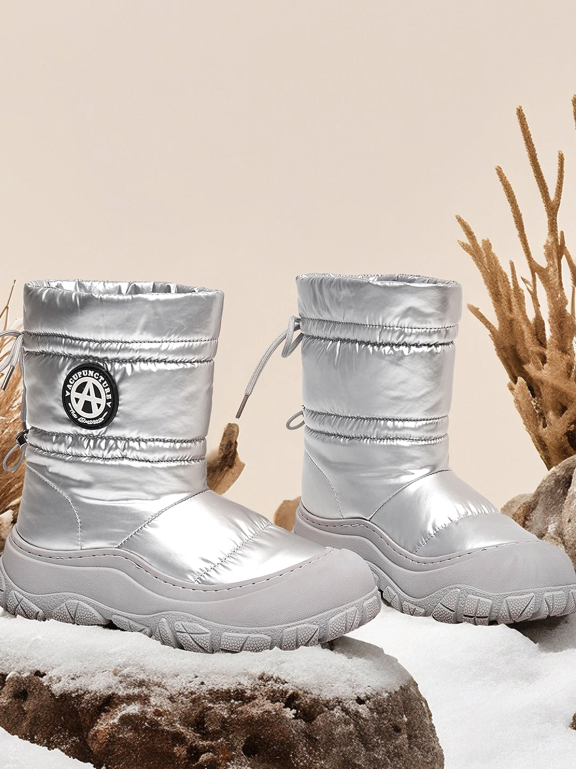 Pair of silver drawstring puffer snow boots displayed in a winter scene with snow, quilted metallic uppers, and grey treaded outsoles
