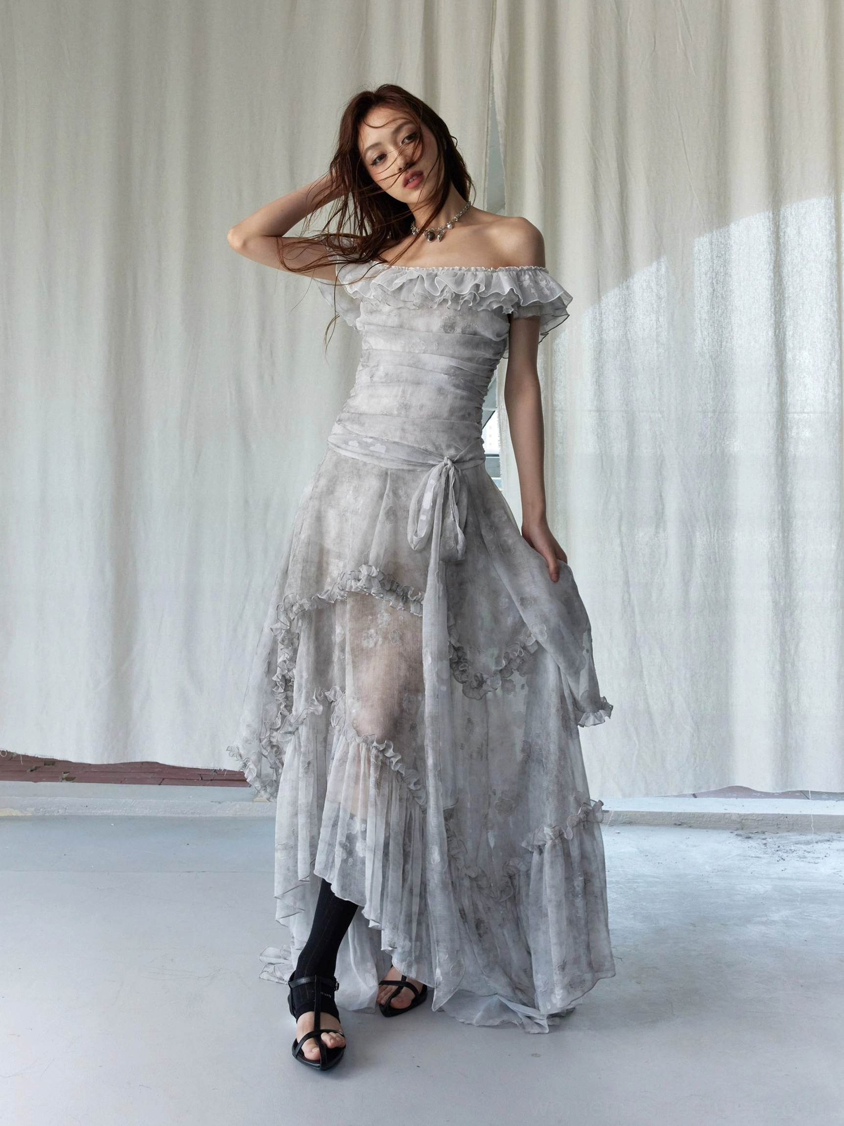 Three-quarter view of silver gray tie-dye off-shoulder ruffled dress with sheer tiered skirt and black crossbody bag
