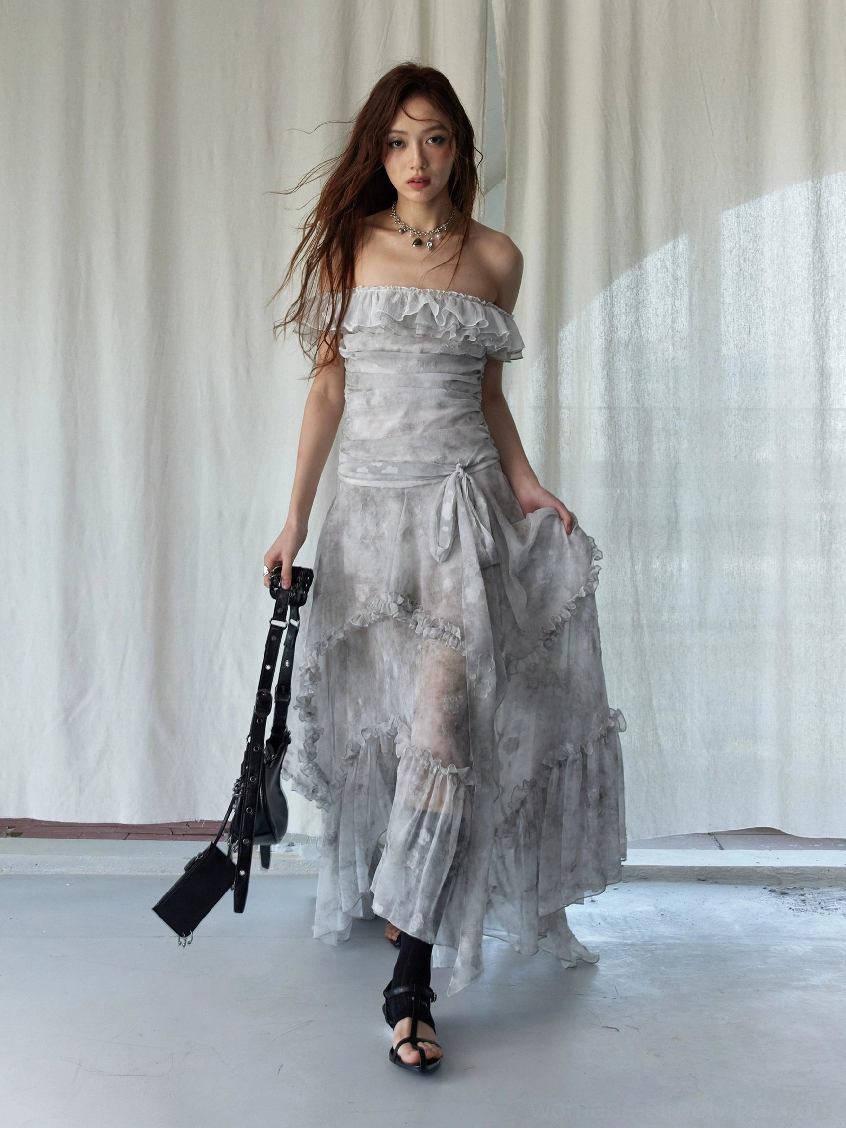 Front view of silver gray tie-dye ruffled maxi dress with asymmetrical hem, ruched waist, black bag, and strappy sandals