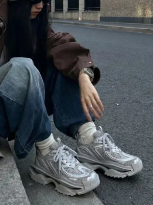 Gray and silver mesh chunky runner sneakers styled with white crew socks, baggy jeans, and a brown jacket in an urban streetwear setting