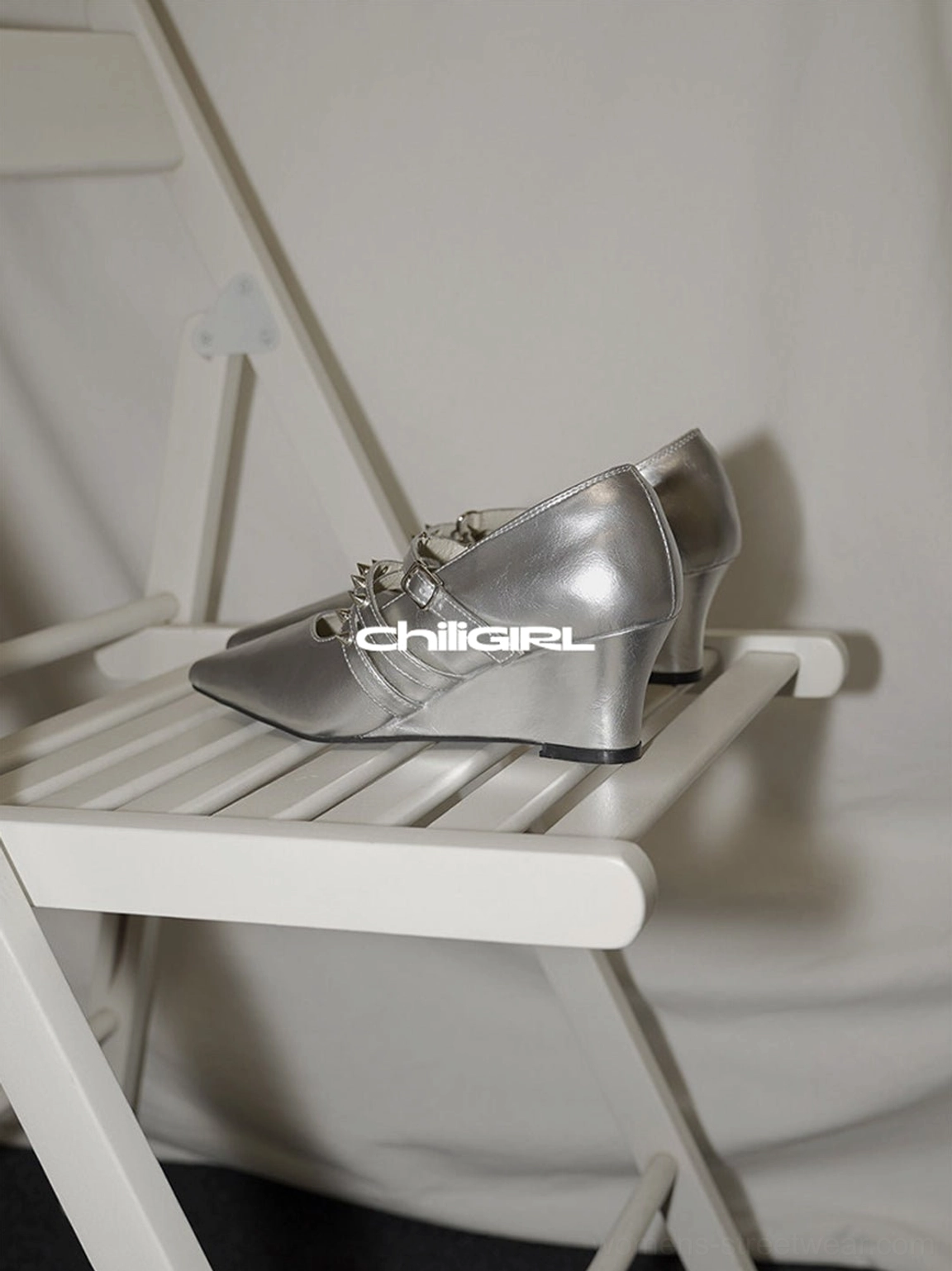 Silver metallic wedge heel pumps with dual adjustable ankle straps and sharp pointed toes on a minimalist chair