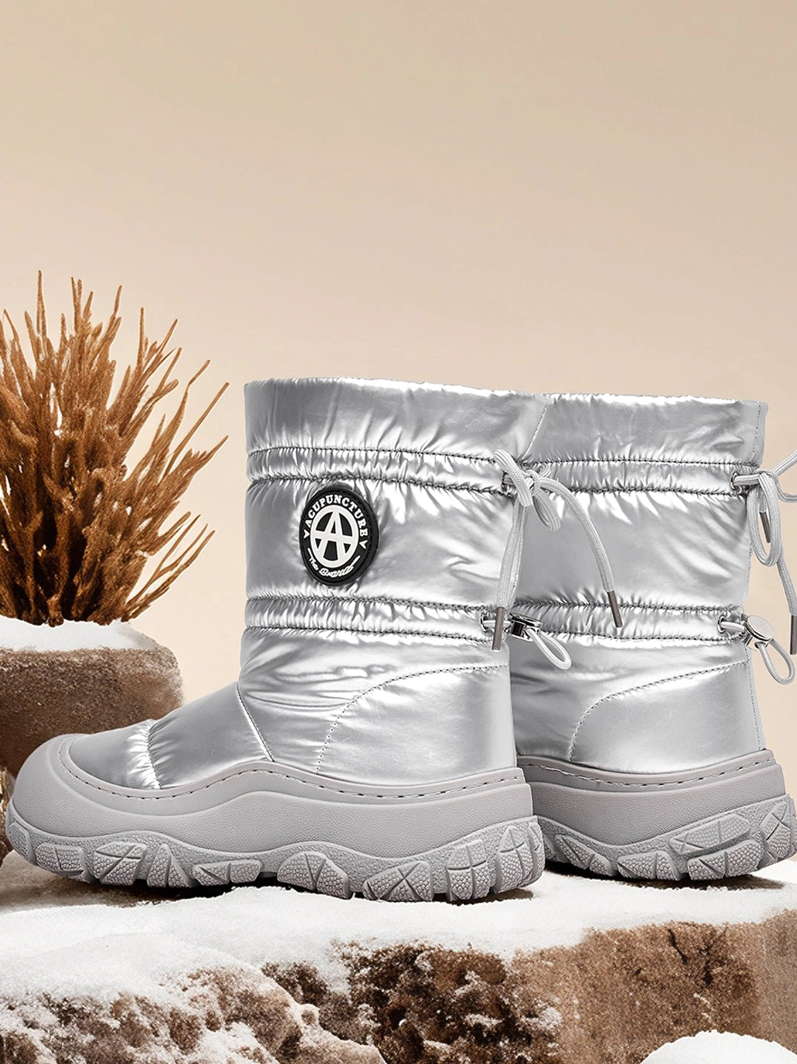 Three-quarter front close-up of silver metallic puffer snow boots in a snowy rustic setting, highlighting the shiny quilted upper