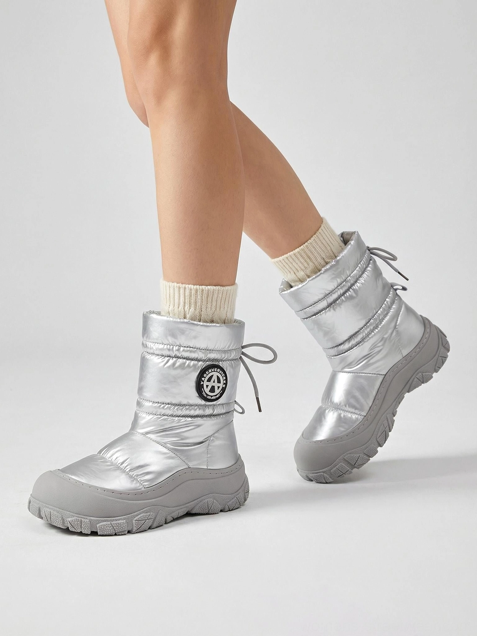 Side view of a model wearing silver metallic puffer snow boots with cream socks, slip-on styling, and chunky grey winter tread soles