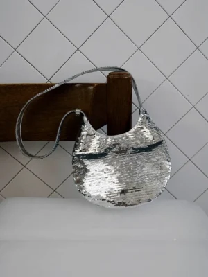 Silver sequin crescent shoulder bag with reflective flip sequins in an indoor lifestyle setting