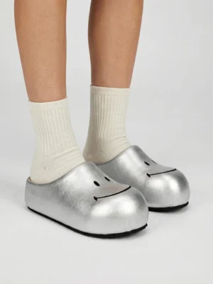 Model wearing silver smiley face slip-on mule sandals with white crew socks, showing the shiny metallic upper and rounded black smile detail from an overhead view