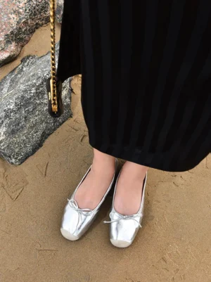Model wearing silver square-toe bow ballet flats with a black striped long skirt on a rocky coastal beach
