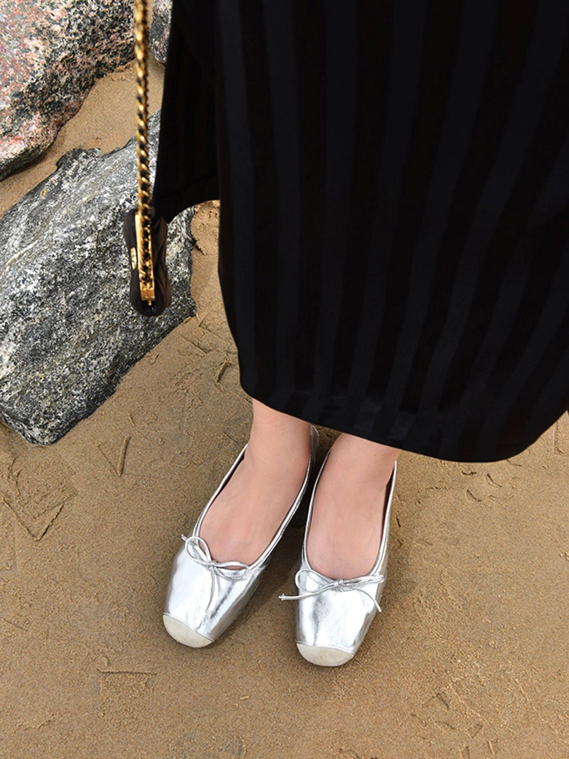Model wearing silver square-toe bow ballet flats with a black striped long skirt on a rocky coastal beach