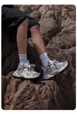 Silver trail running sneakers with rugged rubber outsole worn with black shorts and white crew socks on rocky mountain terrain, showing the chunky gorpcore streetwear silhouette
