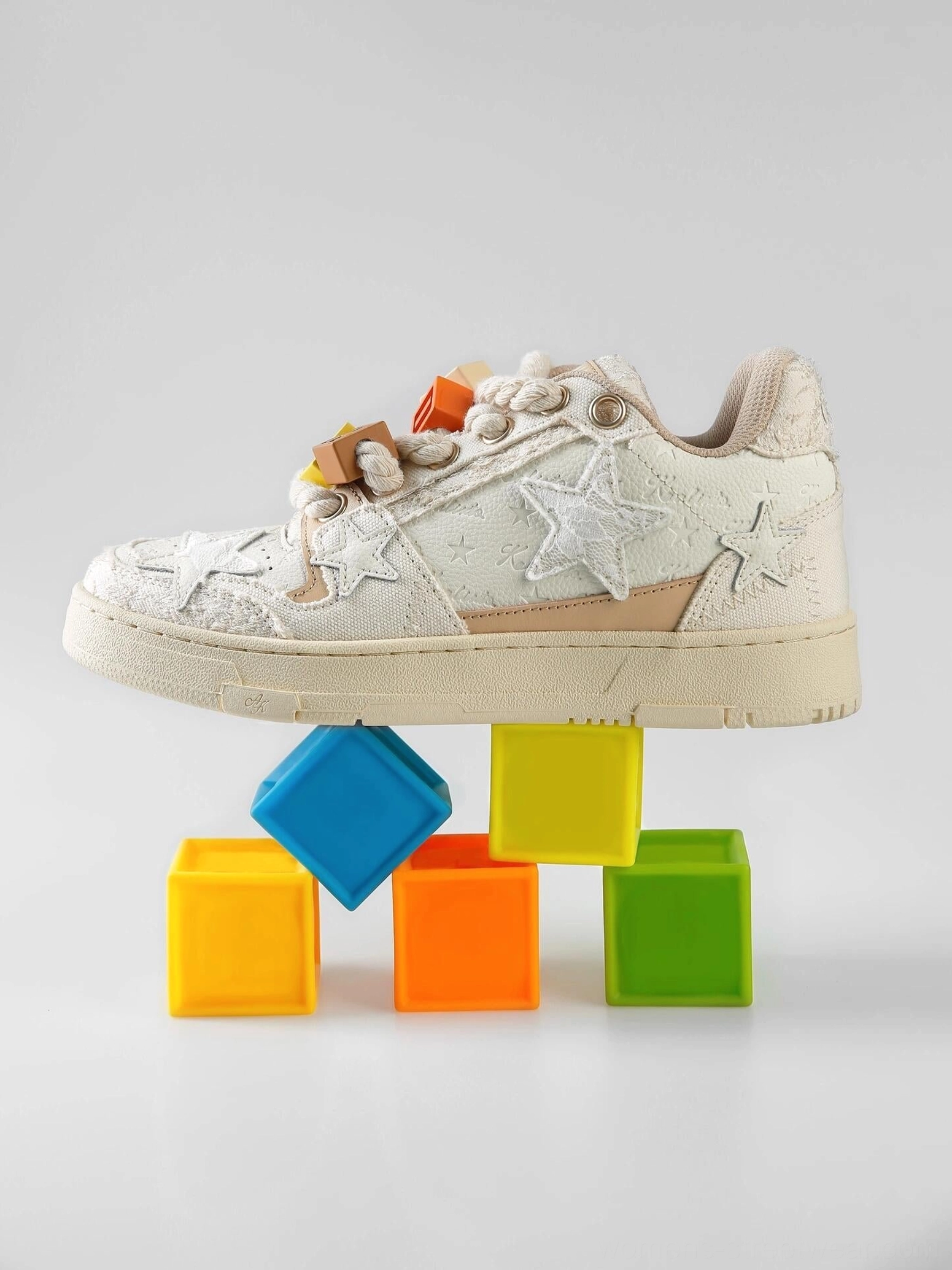 Single beige star sneaker displayed on colorful blocks, showing chunky rope laces, decorative lace charms, textured upper, and off-white rubber sole