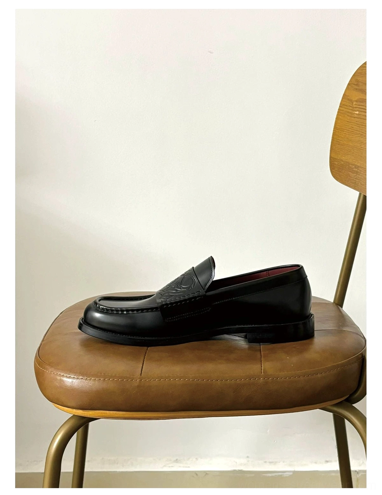 Single black leather loafer displayed on a brown upholstered chair with a classic slip-on silhouette, glossy finish, embossed vamp, and burgundy lining