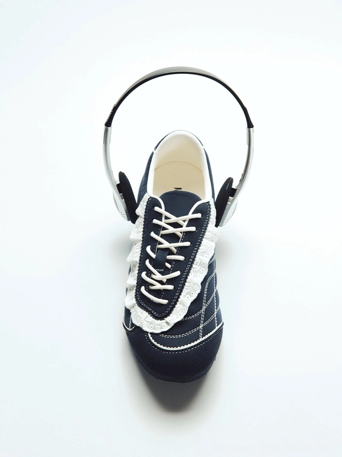 Single dark blue low-profile trainer with white ruffled lace trim and contrast stitching beside vintage headphones