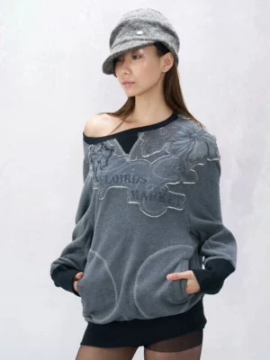 Asian female model wearing a slate blue-green oversized off-shoulder fleece sweatshirt with black ribbed trim, floral appliqué, faded slogan graphic, front pouch pockets, and a fuzzy newsboy bucket hat