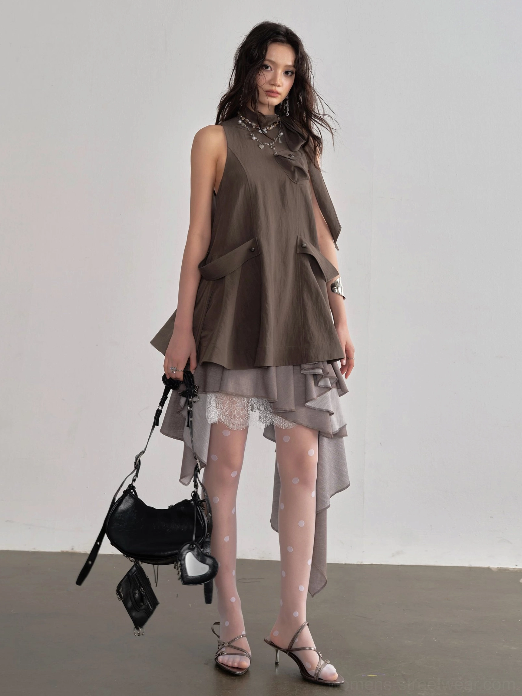 Front view of sleeveless asymmetric cape layer blouse with high neckline, waist buckle details, layered irregular hem, tights, heels, and black shoulder bag