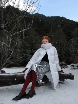 Snow white longline down puffer coat with oversized high collar worn by a woman seated in a snowy mountain setting