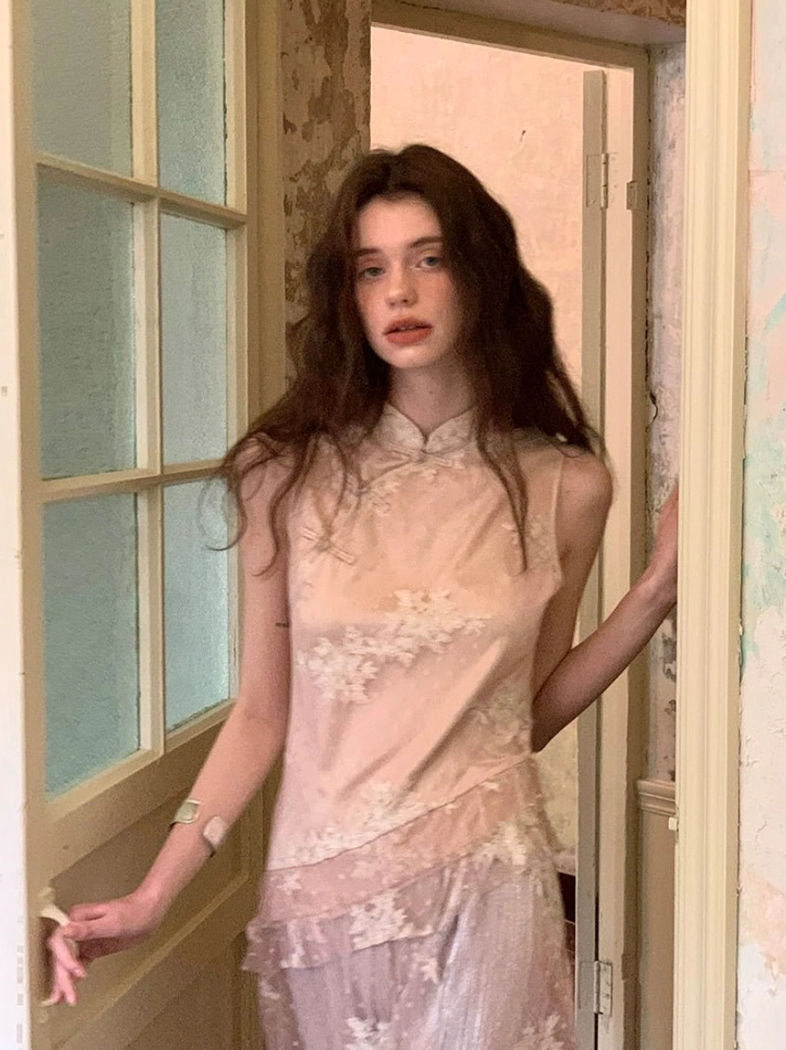 Front view of a soft blush cheongsam-inspired jumpsuit with floral embroidery, sheer asymmetric layering, and refined silver jewelry