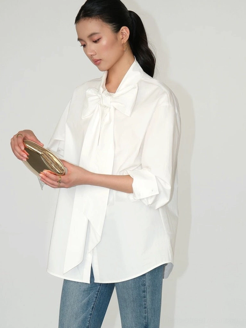 Asian female model wearing a solid white oversized cotton bow blouse with rolled cuffs and blue jeans in a minimalist Korean fashion look