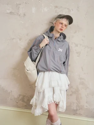 Front view of gray sporty hooded windbreaker jacket and tiered lace midi skirt set with chest bow embroidery, contrast piping, baseball cap, crossbody bag, and ruffled lace socks