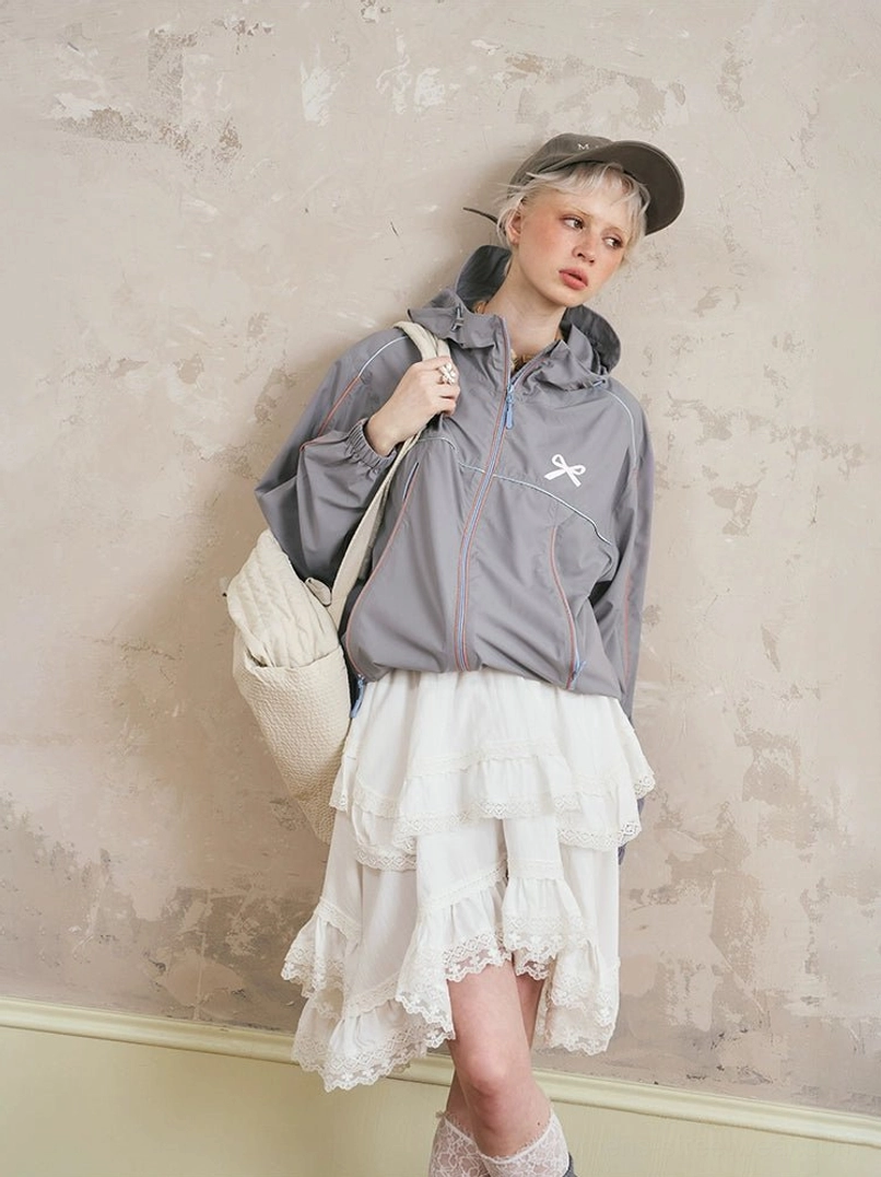 Front view of gray sporty hooded windbreaker jacket and tiered lace midi skirt set with chest bow embroidery, contrast piping, baseball cap, crossbody bag, and ruffled lace socks