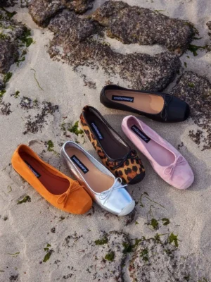 Women’s square-toe bow ballet flats in multiple colorful velvet-look finishes displayed on a sandy coastal beach
