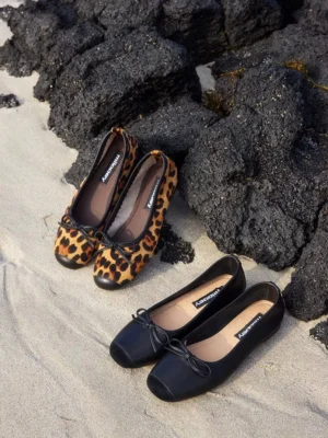 Leopard print and black square-toe bow ballet flats for women arranged side by side on sand near dark rocks