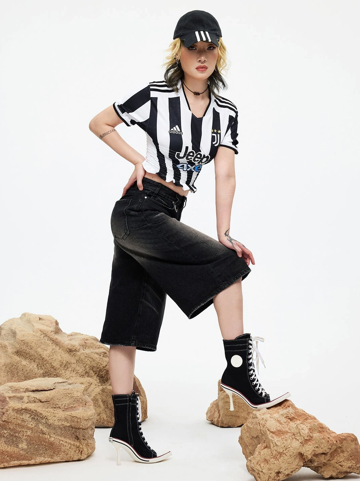 Standing female model front three-quarter view wearing white canvas stiletto boots paired with striped jersey, denim culottes and baseball cap on brown rock decorated studio set