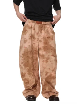 Front low angle view of apricot beige star etched desert satin wide leg streetwear trousers with rust starburst pattern, elastic waistband, oversized relaxed fit, and stacked floor pooling silhouette