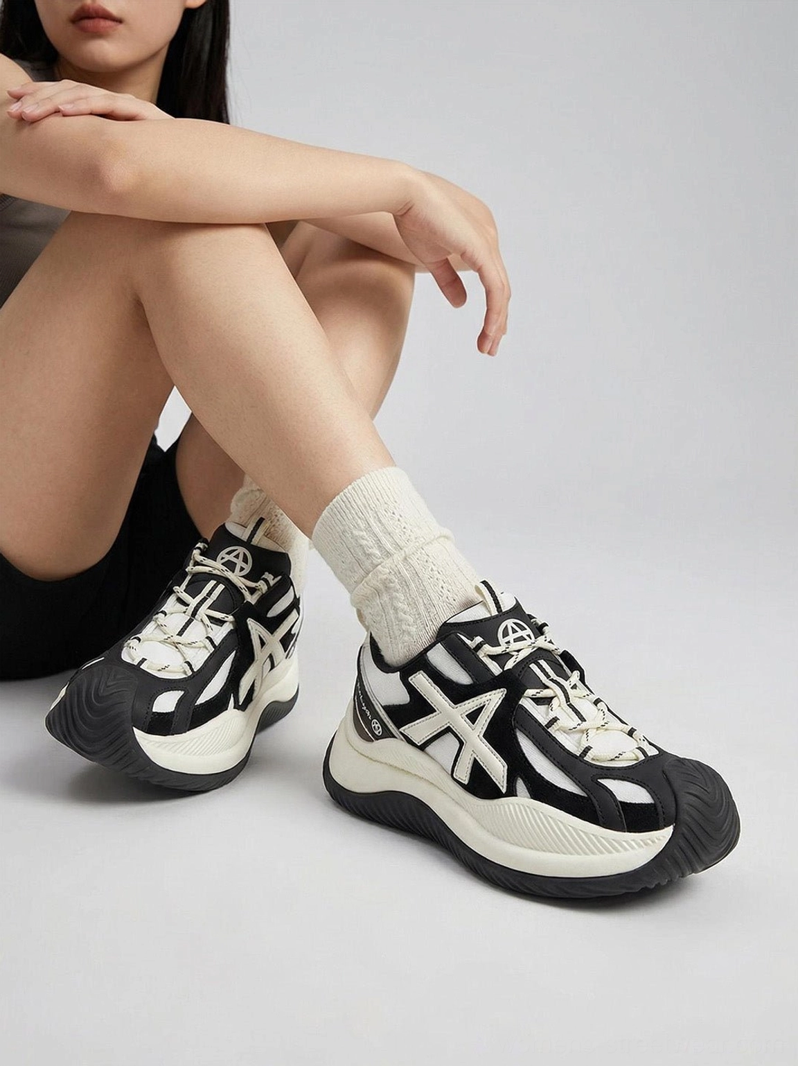 Seated model view of chunky sneakers highlighting panda-style black white color match and decorative cross toe cap detail