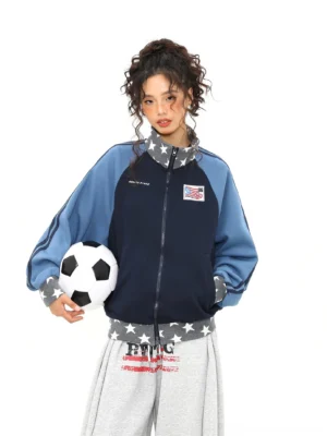 Female model wearing a navy and blue star print colorblock varsity track jacket while holding a soccer ball in a white studio