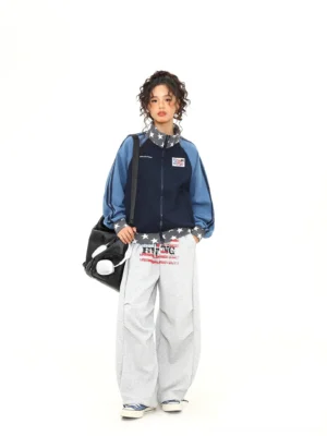 Full body view of a female model wearing a relaxed colorblock varsity track jacket with grey wide leg sweatpants and a black shoulder bag