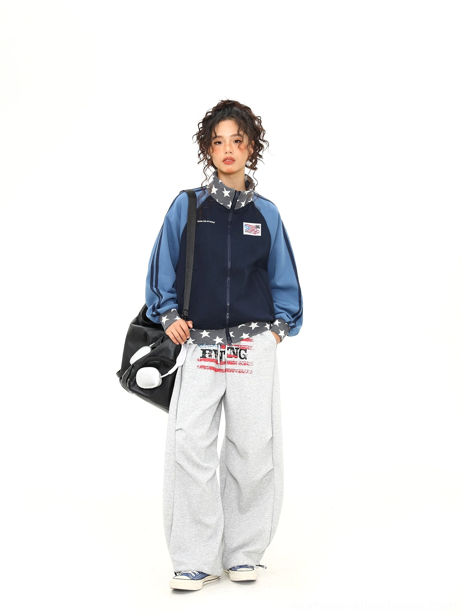 Full body view of a female model wearing a relaxed colorblock varsity track jacket with grey wide leg sweatpants and a black shoulder bag