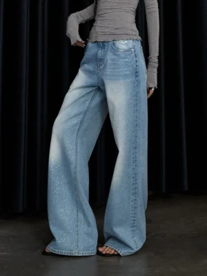 Side low-angle view of washed blue stardust glitter wide-leg jeans styled with a gray cropped long-sleeve top in a streetwear studio look