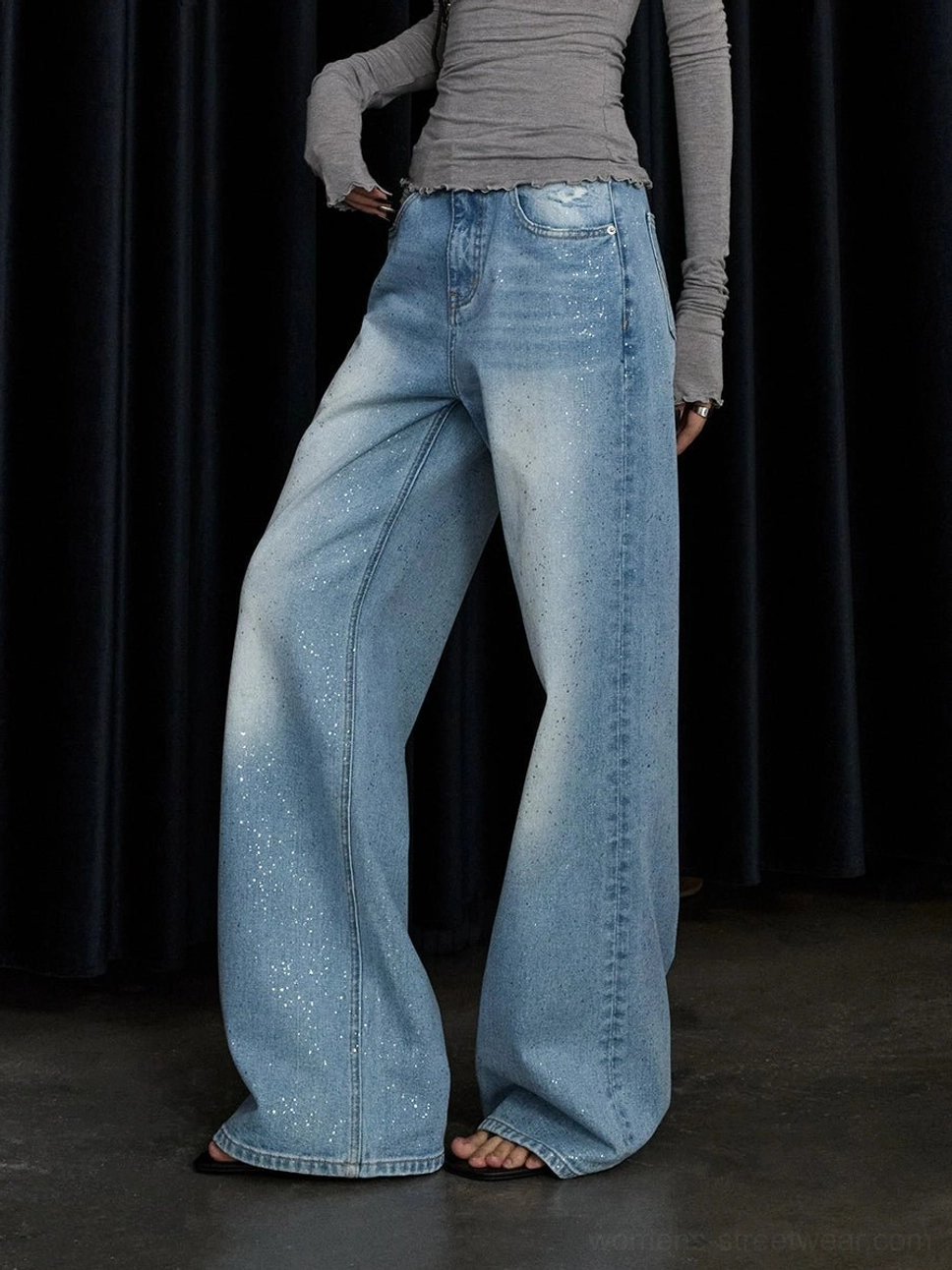 Side low-angle view of washed blue stardust glitter wide-leg jeans styled with a gray cropped long-sleeve top in a streetwear studio look