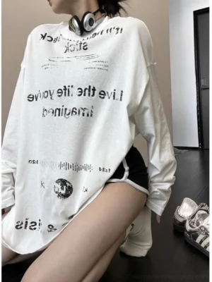 White oversized long sleeve graphic tee with inverted black retro typography and planet print, styled with black athletic shorts in an indoor hallway
