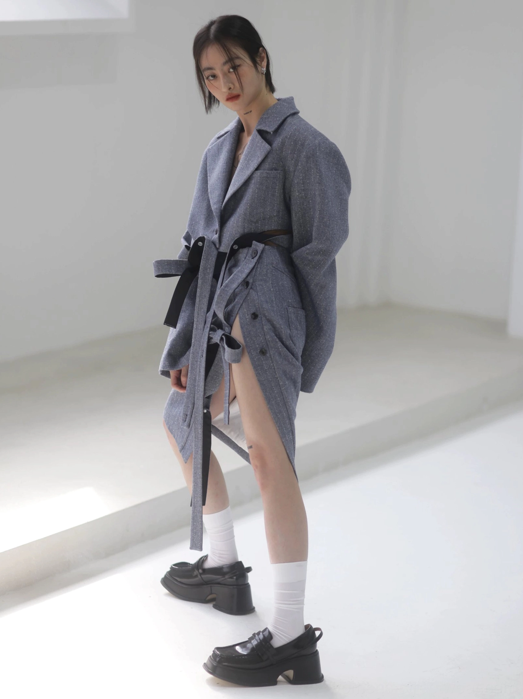 Three-quarter view of a steel blue herringbone midi pencil skirt with side slit and ribbon fastening styled with a tailored coat and waist belt
