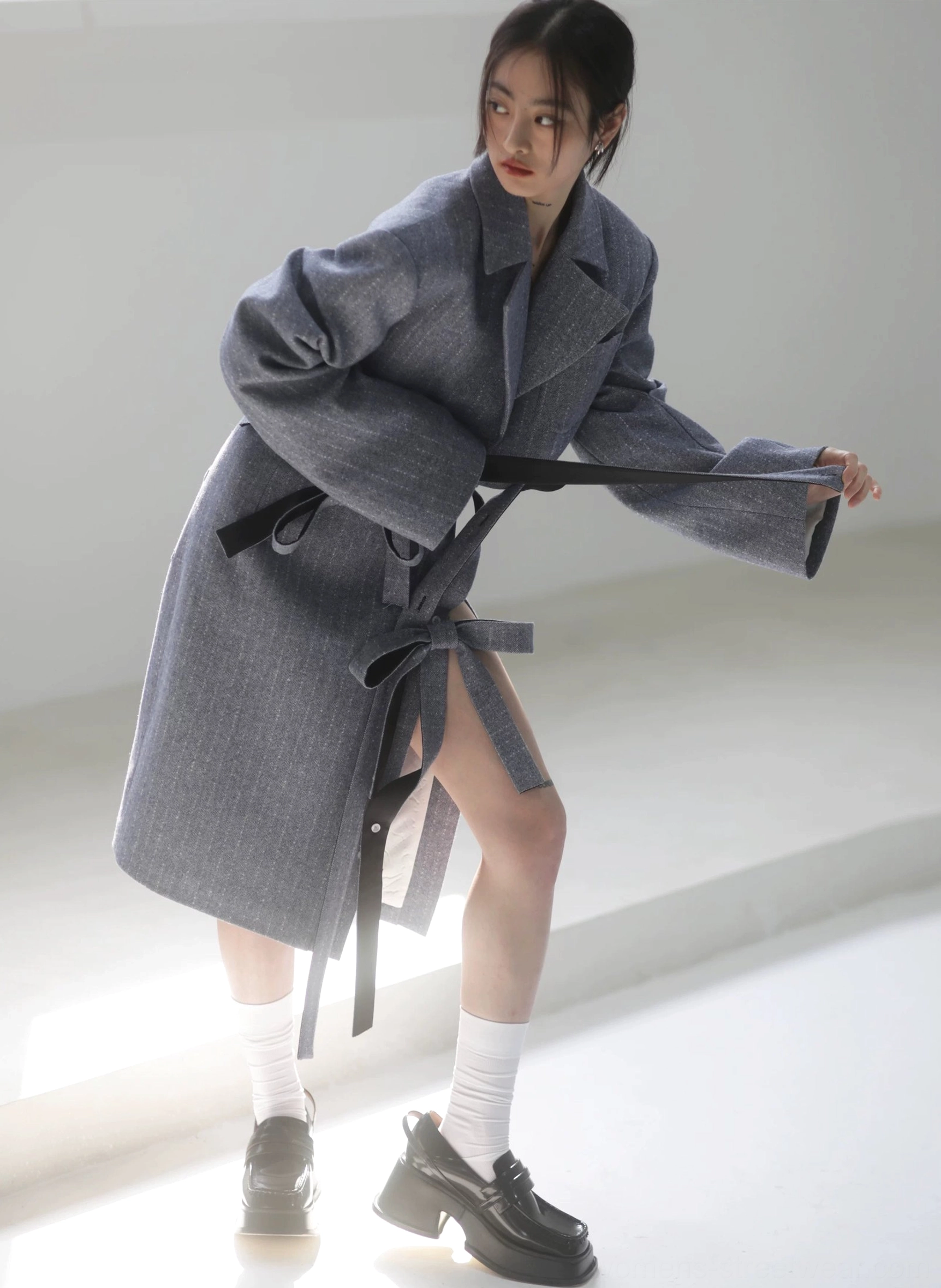 Dynamic side view of a steel blue herringbone pencil midi skirt with adjustable ribbon belt and side slit styled under a tailored long coat