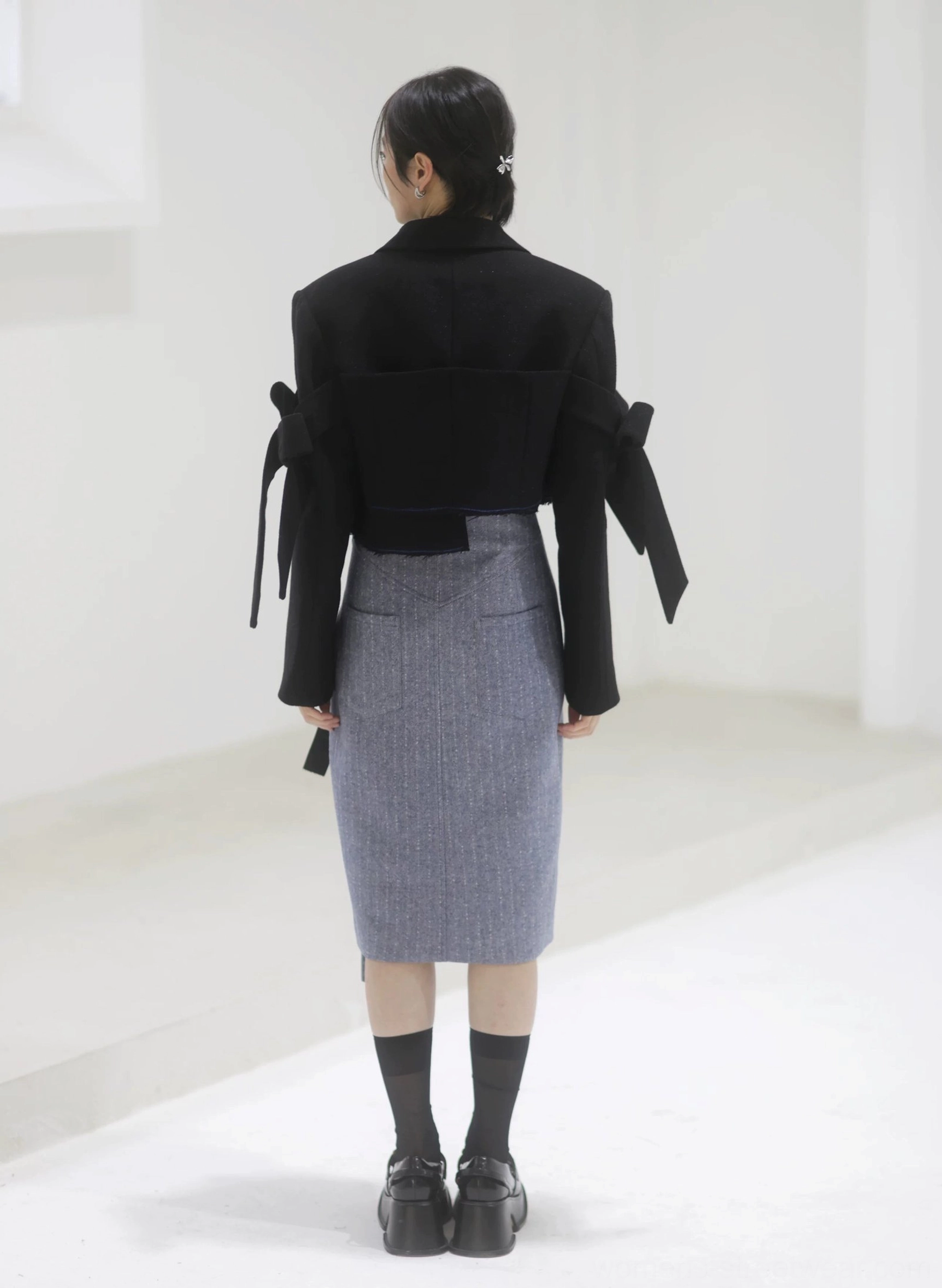 Full back view of a steel blue herringbone pencil midi skirt with a straight midi silhouette styled with a cropped black blazer and bow sleeve details