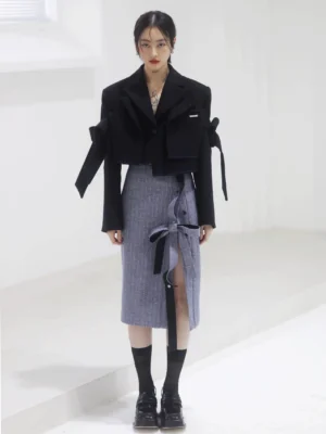 Steel blue grey herringbone pencil midi skirt styled with a cropped black blazer in a minimalist studio, showing the high waist, tailored column silhouette, side slit, and ribbon tie closure